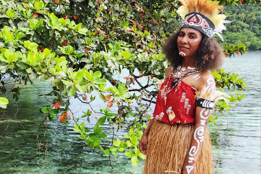 Person in traditional attire standing on a wooden platform by a body of water with greenery.